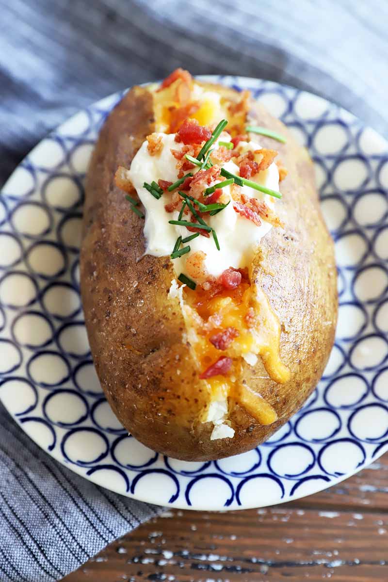 Vertical image of a single potato stuffed with cheese, bacon, sour cream, and fresh herbs on top.