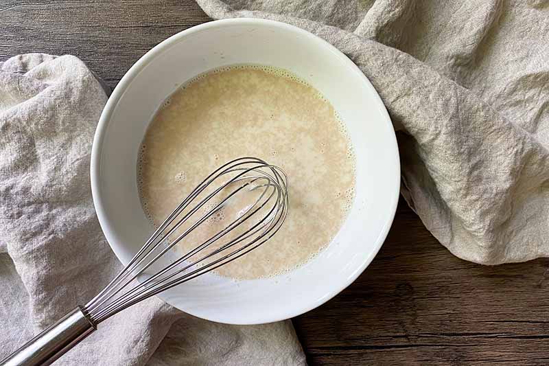 Horizontal image of whisking together yeast and milk in a white bowl.