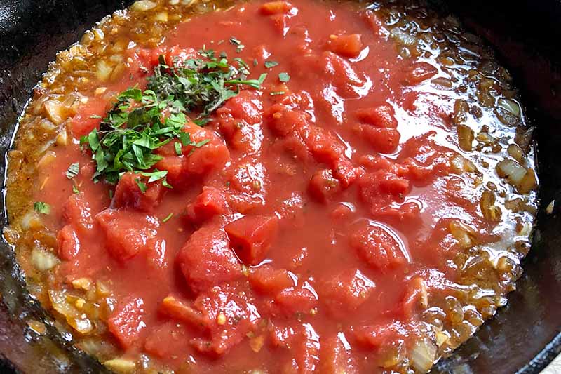 Horizontal image of stewed tomatoes with a small pile of chopped herbs in a skillet.