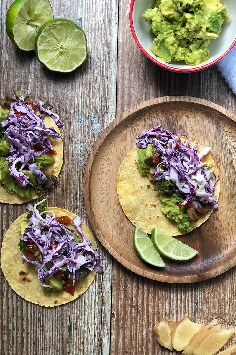 Vertical top-down image of corn tortillas topped with cooked vegetables and red cabbage slaw next to limes and smashed avocados on a wooden table.
