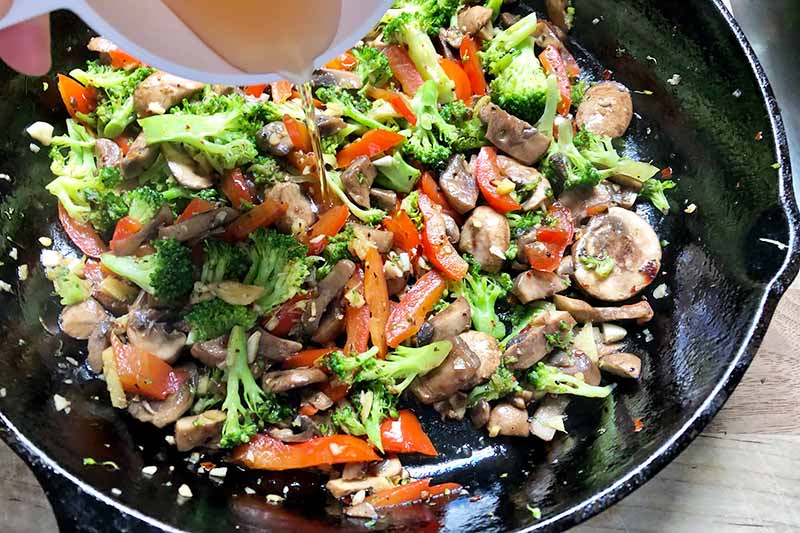 Horizontal image of broccoli, peppers, and mushrooms cooking in a pan with broth being poured on top of them.