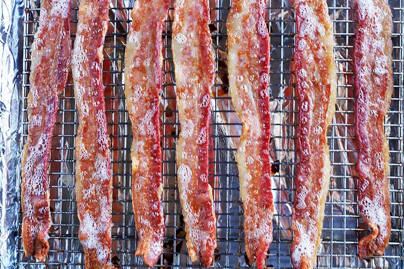 Horizontal image of cooked slices of meat on a cooling rack lined with aluminum foil to catch the dripping fat.