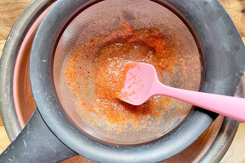 Horizontal image of a pink spatula pushing a thick orange liquid through a strainer.