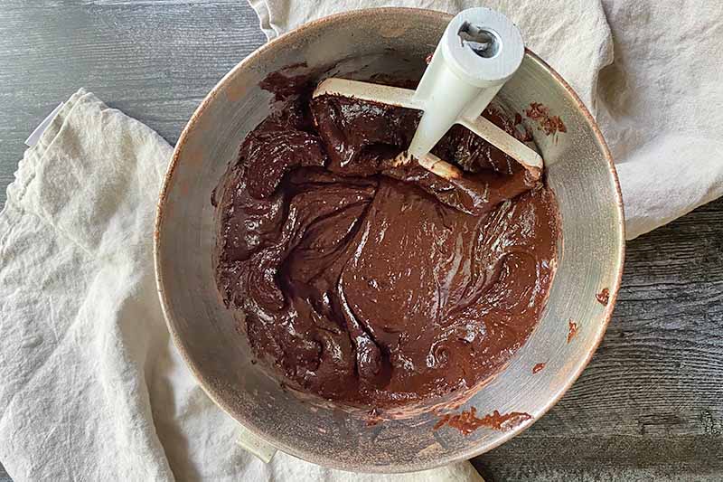 Horizontal image of a thick brown batter in a bowl with a paddle attachment.