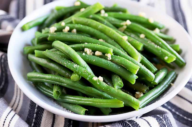 Horizontal image of seasoned cooked green beans in a white bowl on a gray and white towel.