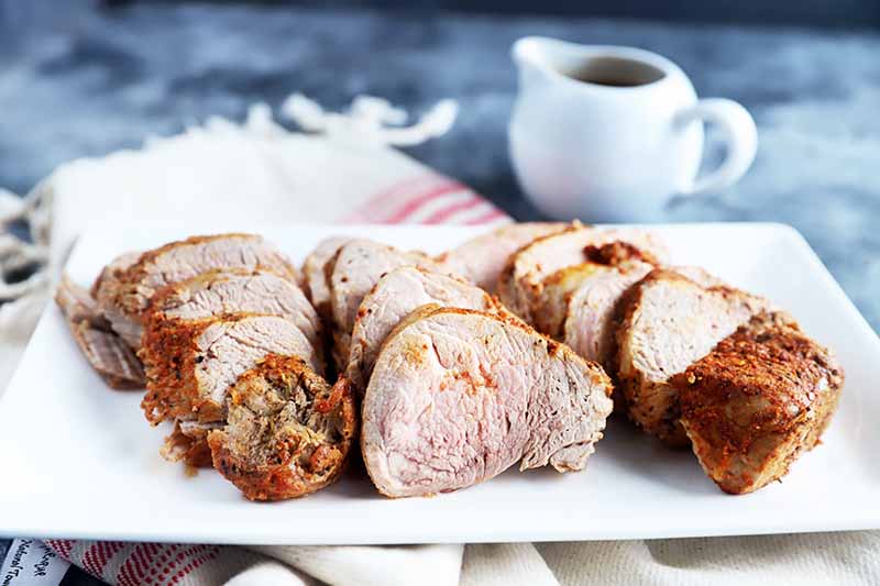 Horizontal image of slices of cooked seasoned meat on a white plate next to a cup of gravy and a towel.