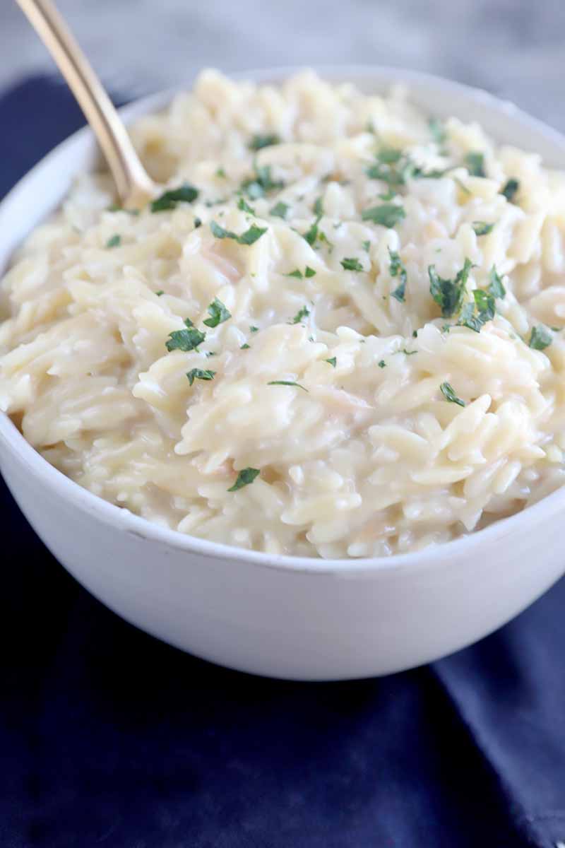 Vertical image of a white bowl filled with a creamy and cheesy pasta topped with chopped fresh herbs with a spoon inserted into the bowl.