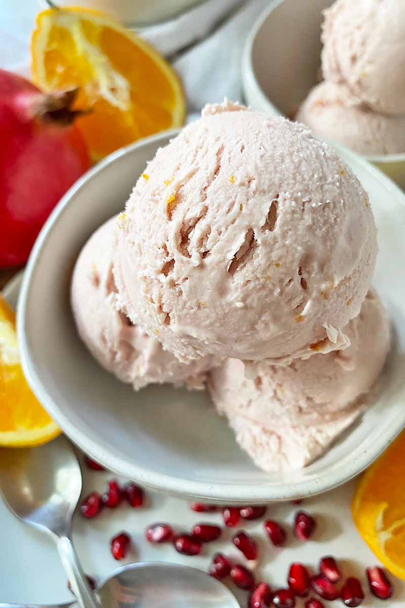Vertical image of a white bowl filled with three scoops of a light pink frozen dessert next to fruit and spoons.