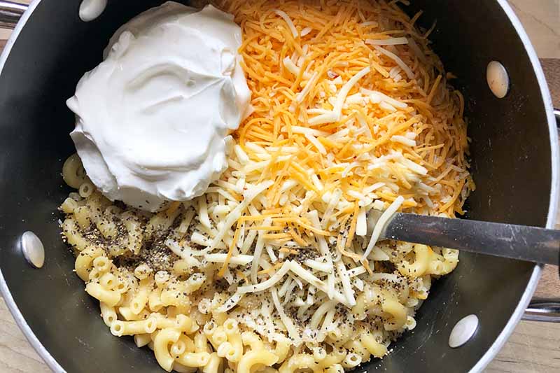 Horizontal image of a dollop of sour cream, shredded cheese, and pasta in a pot stirred by a spoon.