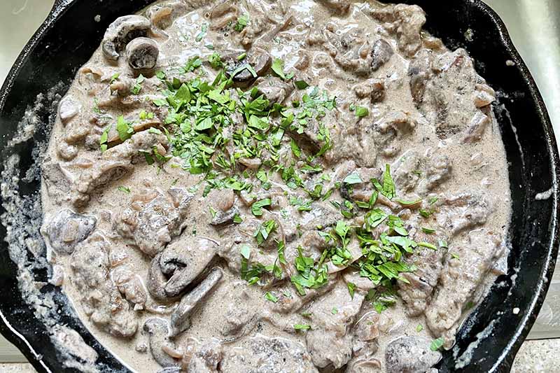 Horizontal image of cooked meat in a cream sauce with herb garnish in a skillet.