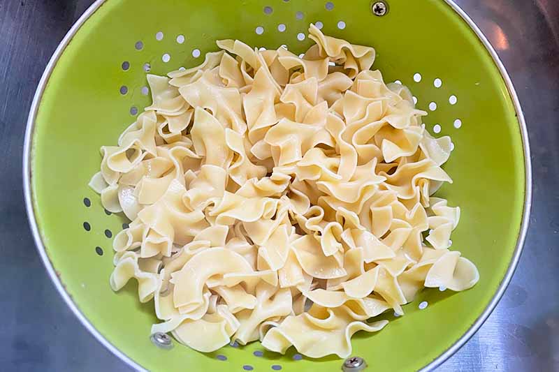 Horizontal image of a green colander with cooked egg noodles.