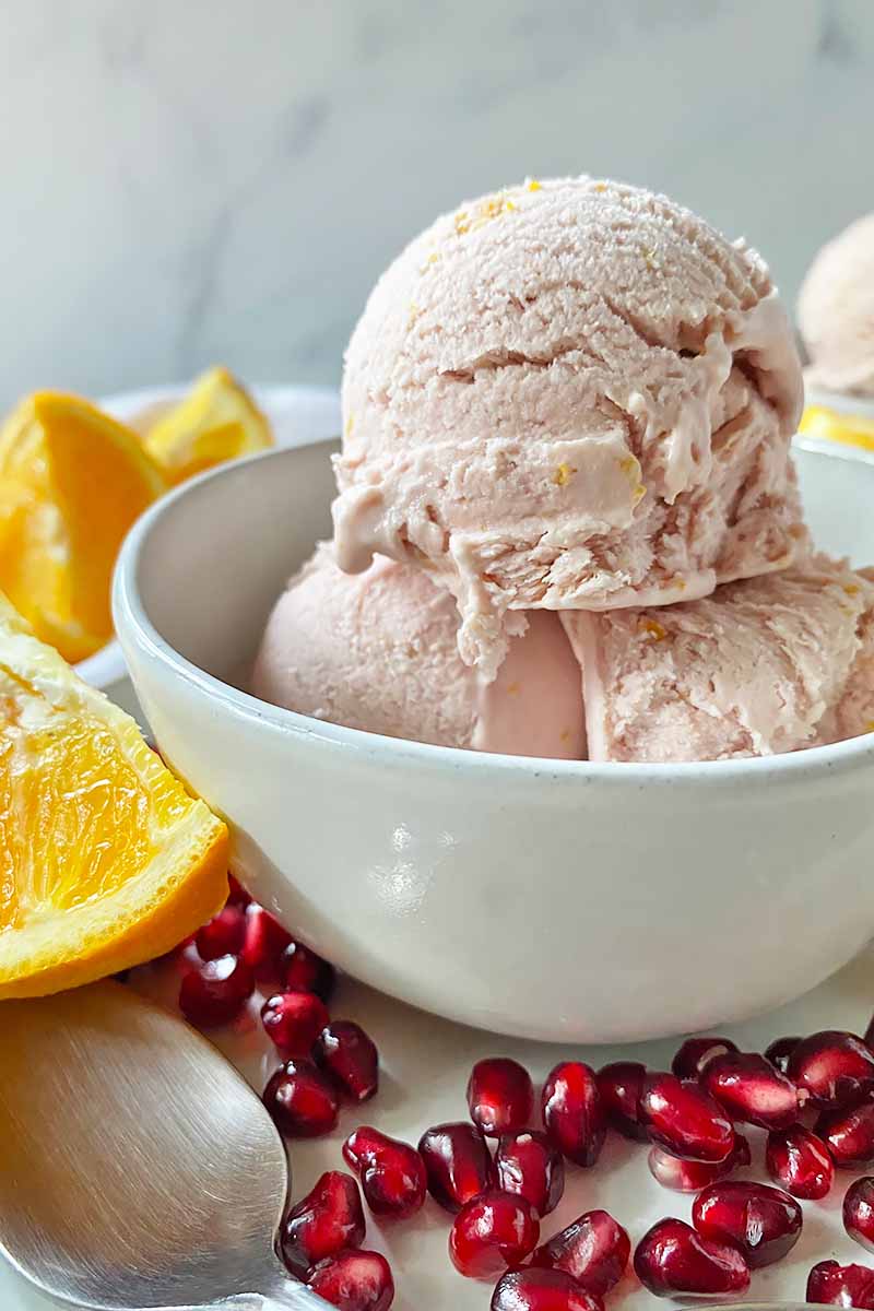 Vertical image of a small white bowl with three scoops of a light pink frozen dessert next to slices of citrus and fruit.