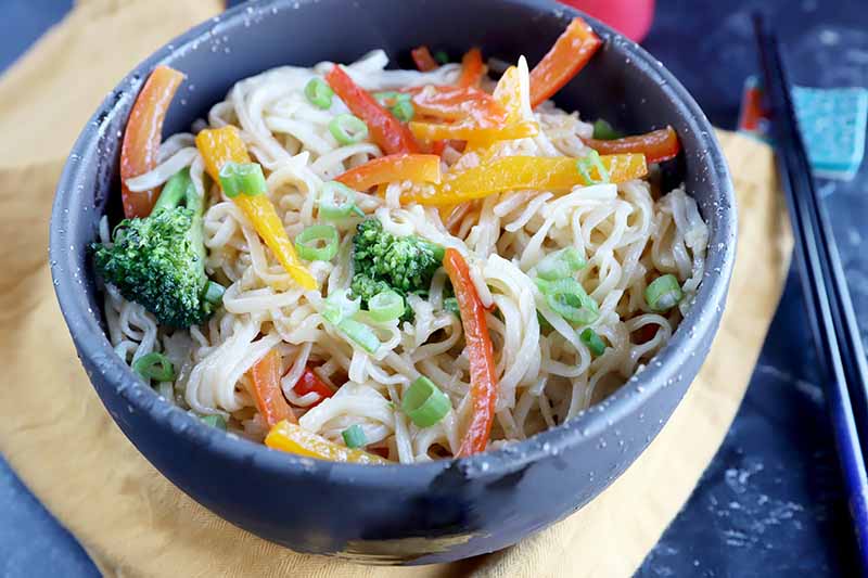 Horizontal image of rice noodles mixed with assorted vegetables in a gray bowl on a yellow towel.