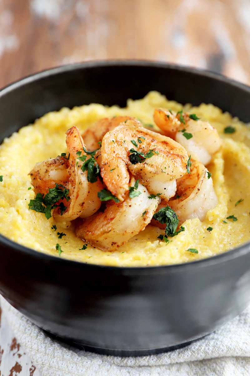 Vertical image of a black bowl full of creamy cornmeal topped with seasoned shrimp on a white towel.