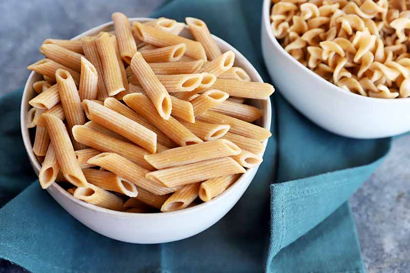 Horizontal image of a bowl of penne and a bowl of fusilli on a blue towel.