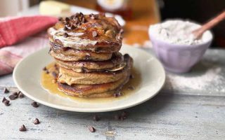 Horizontal image of a tall stack of pancakes with maple syrup and chocolate chips next to a bowl of flour and a red plaid towel.