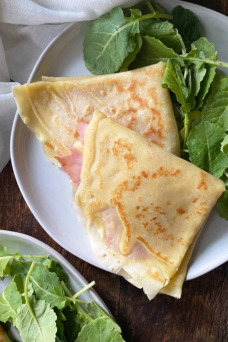 Vertical top-down image of two ham and cheese breakfast flatbreads next to a bed of greens on a white plate.