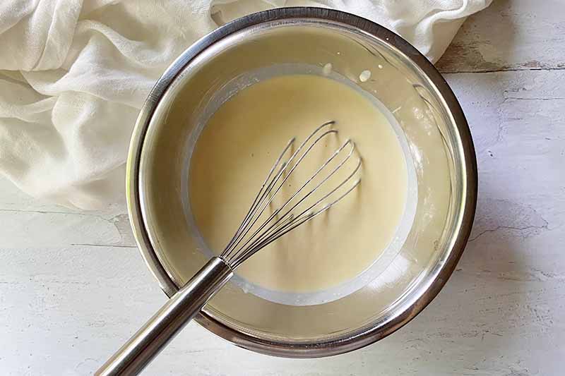 Horizontal image of a thin light yellow batter with a whisk in a bowl.