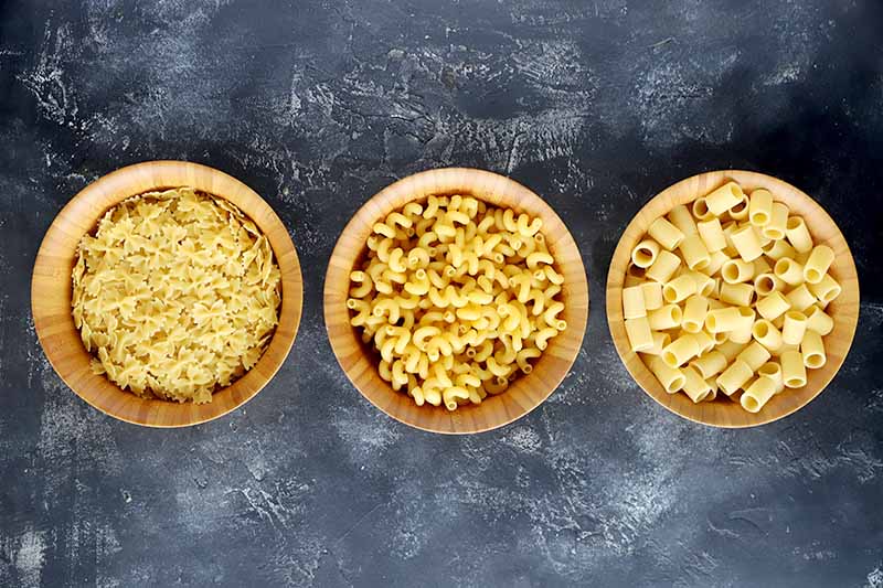 Horizontal image of farfalle, fusilli, and rigatoni in wooden bowls on a gray surface. 