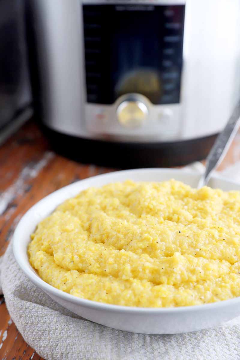 Vertical image of a white bowl full of creamy grits with a spoon in front of a kitchen appliance.