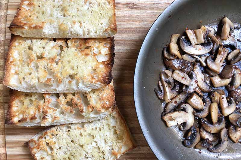 Horizontal image of toasted slices of bread and a pan with cooked sliced mushrooms.