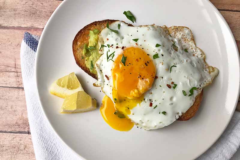 Horizontal image of a sunny-side up egg on toast next to lemon slices on a white plate.