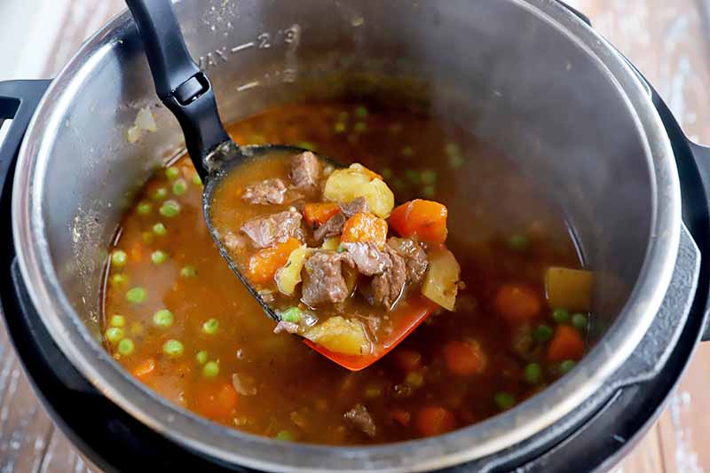 Horizontal image of a spoon holding some soup with chunks of meat and vegetables over a steaming pot.