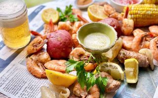 Horizontal image of shrimp, sausage, lemons, and assorted vegetables on newspaper next to dip and beer.