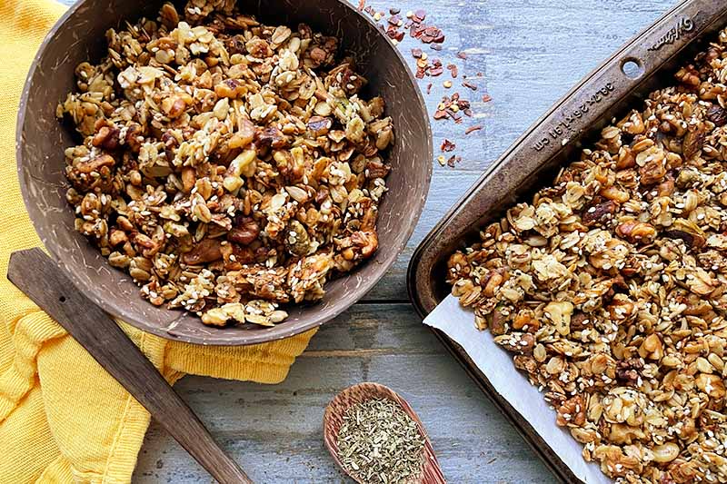 Horizontal top-down image of a wooden bowl and a baking sheet with clusters of oats and nuts with wooden spoons filled with spices.