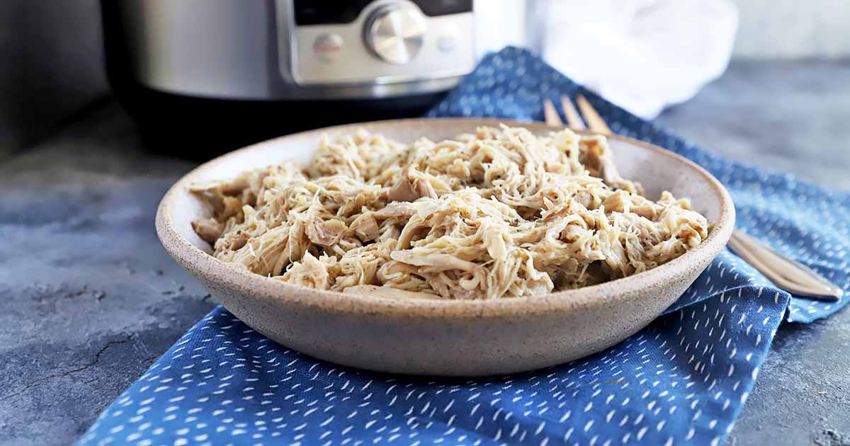 Horizontal image of a beige ceramic bowl of shredded cooked poultry, on a blue folded cloth napkin with white speckles, with a fork on a blue marble countertop, with an Instant Pot in the background.