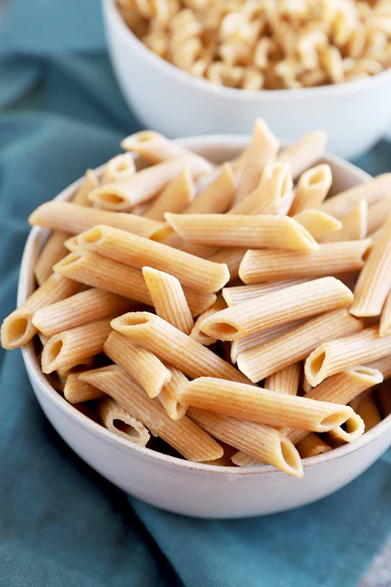 Vertical image of a bowl of penne and a bowl of fusilli over a blue towel.