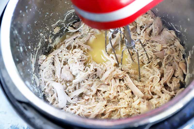 Horizontal image of a hand mixer pulling apart juicy cooked poultry in a pot.