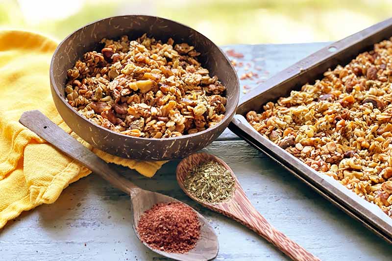 Horizontal image of a wooden bowl and a baking sheet filled with an oat, nut, and seed mixture next to a yellow towel and wooden spoons filled with spices.