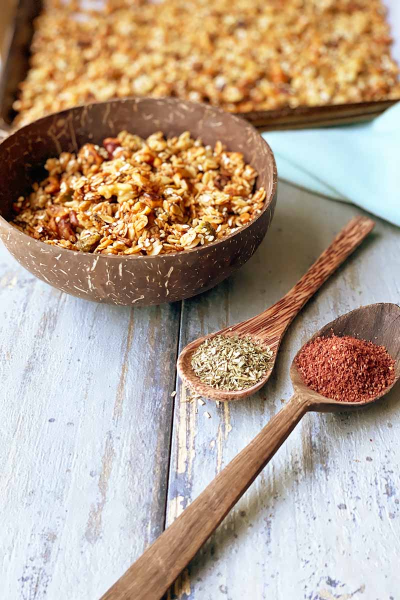 Vertical image of a wooden bowl filled with a mix of oats and nuts on a wooden table next to a baking sheet, blue towel, and wooden spoons filled with ground spices.