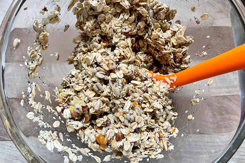 Horizontal image of mixing oats, nuts, and seeds with an orange spatula in a glass bowl.
