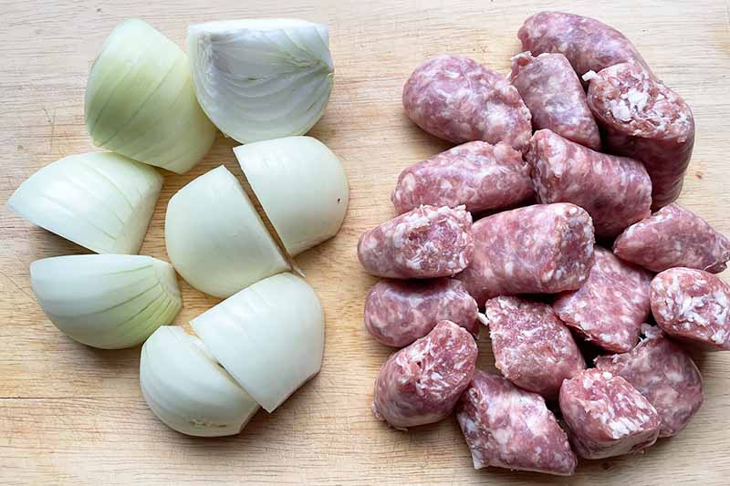 Horizontal image of quartered onions and sliced sausage on a cutting board.