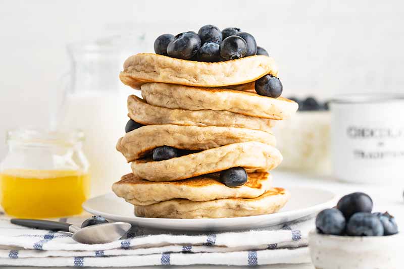 Horizontal image of a stack of pancakes with blueberries on a towel next to orange juice and milk.