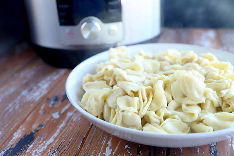Horizontal image of a white bowl of stuffed pasta in front of a kitchen appliance on a wooden surface.