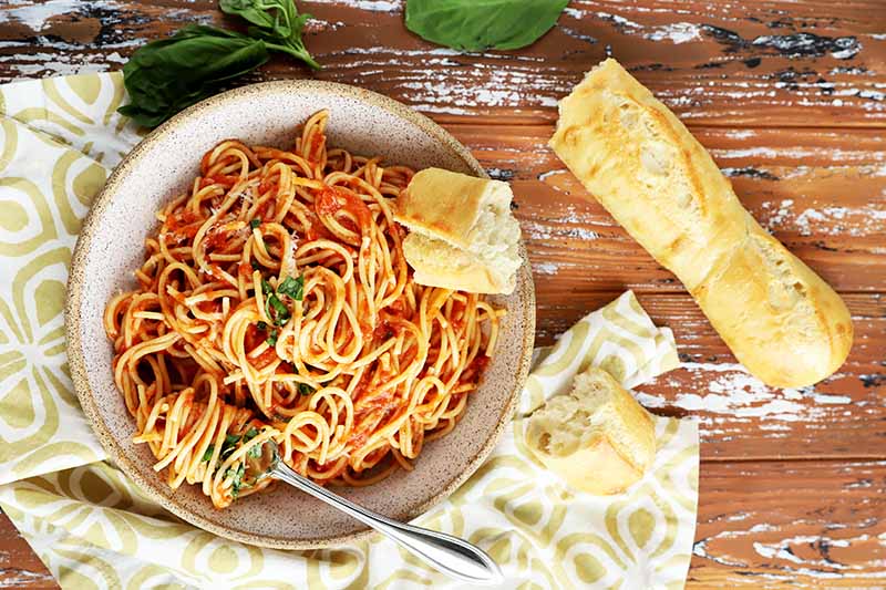 Horizontal image of spaghetti and tomato sauce in a bowl next to a yellow towel and part of a baguette.