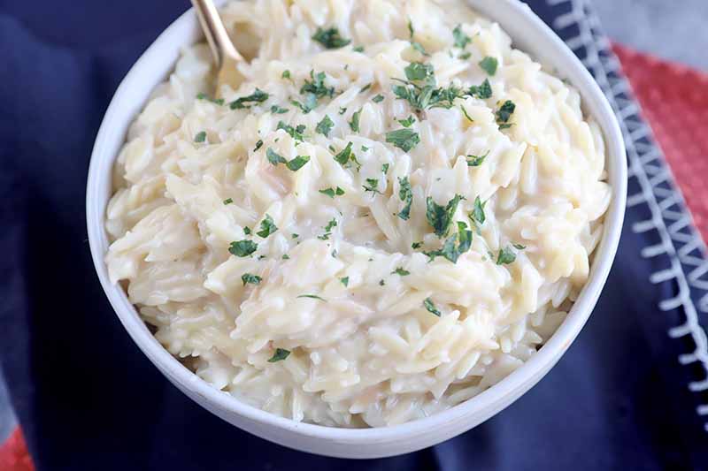 Horizontal image of a creamy orzo recipe in a white bowl garnished with chopped fresh herbs.