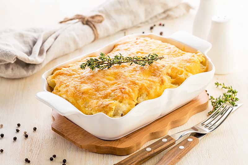 Horizontal image of a cheesy casserole in a white baking dish next to a tan towel and silverware.