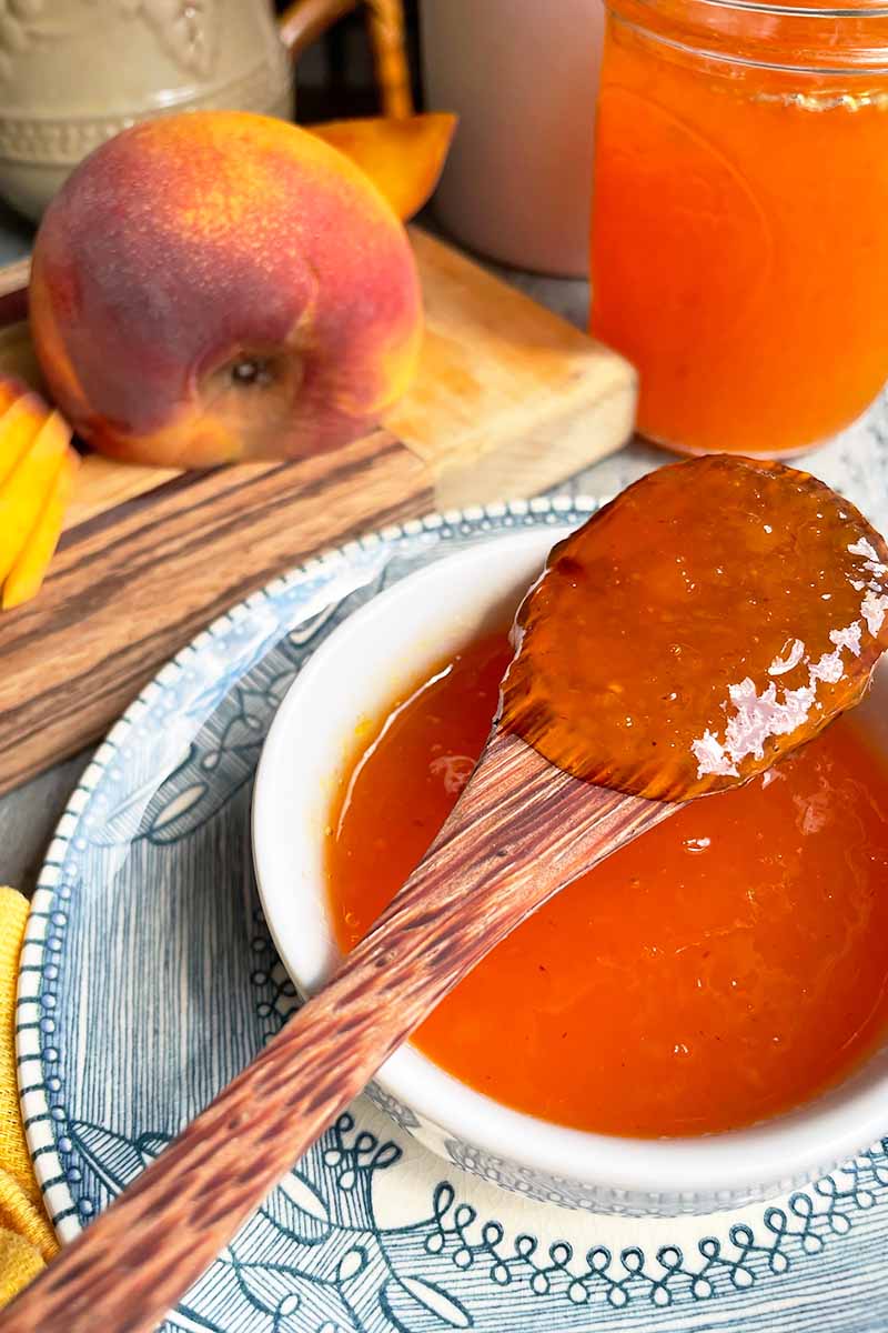 Vertical image of a wooden spoon over a bowl holding an orange-colored jelly in front of fresh fruit and a jar on a wooden board.