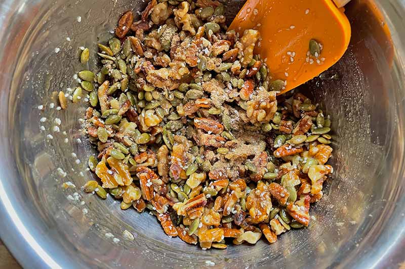 Horizontal image of an orange spatula mixing together assort4ed nuts and seeds in a metal bowl.