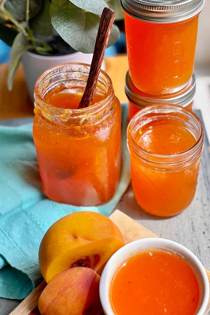 Vertical image of jars full of an orange-colored spread on a blue towel next to a bowl and sliced fresh fruit.