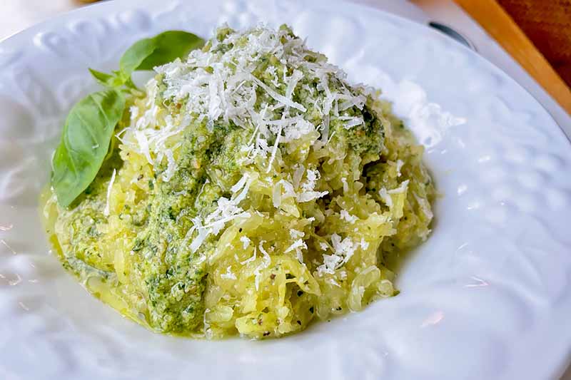 Horizontal image of a pile of shredded yellow vegetables in a light green sauce with herb and cheese garnishes on a white plate.