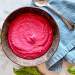 Horizontal image of a wooden bowl filled with a bright pink thick spread next to a blue towel with a metal spoon surrounded by fresh herbs.