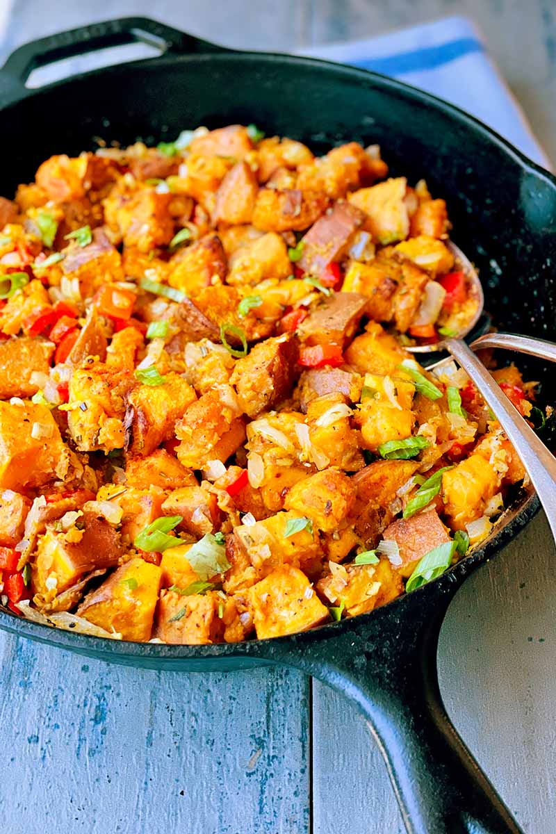 Vertical image of a cast iron skillet filled with cubed orange vegetables with seasonings on a wooden surface.