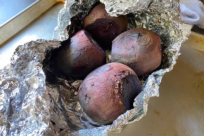 Horizontal image of whole cooked purple root vegetables in an aluminum foil package on a baking sheet.
