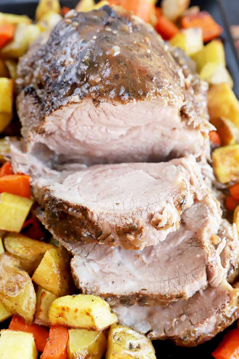 Vertical image of a large piece of meat with slices in a serving dish surrounded by diced potatoes and carrots.