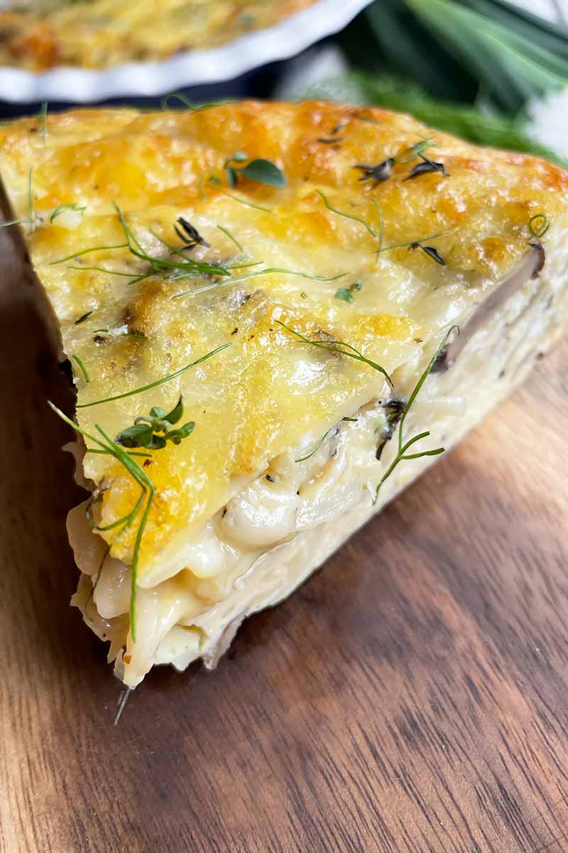 Vertical close-up image of a slice of frittata with herbs and vegetables on a wooden plate.